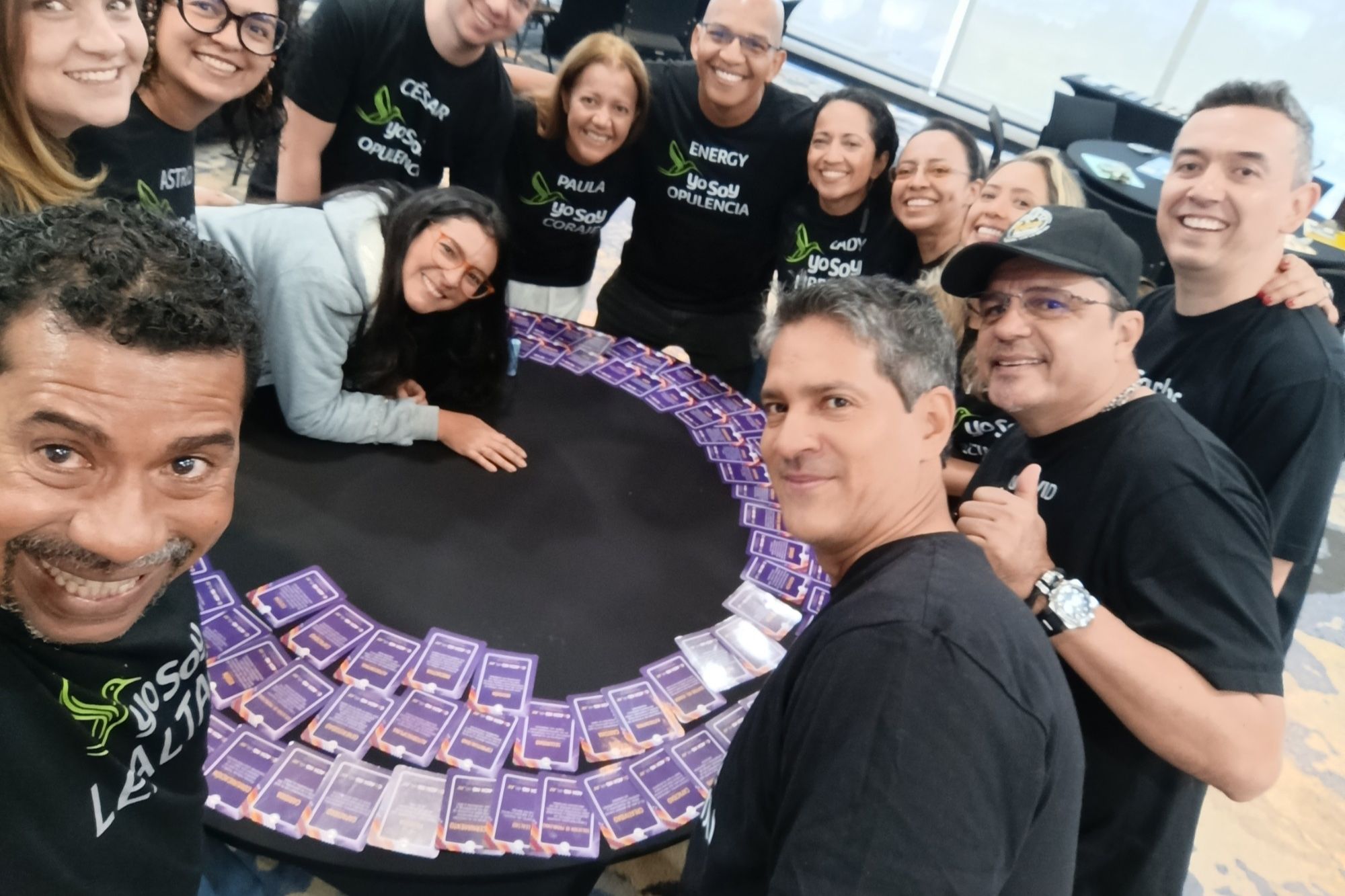 Selfie de un grupo de facilitadores con camisetas Yo Soy alrededor de una mesa cubierta de cartas moradas.
