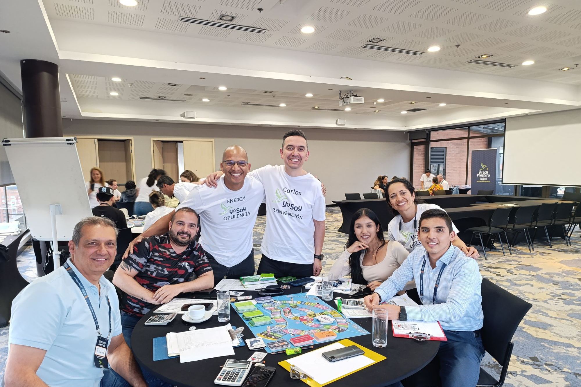 Aprendices sonrientes alrededor del tablero del simulador financiero, con dos facilitadores de camiseta Yo Soy abrazados detrás.