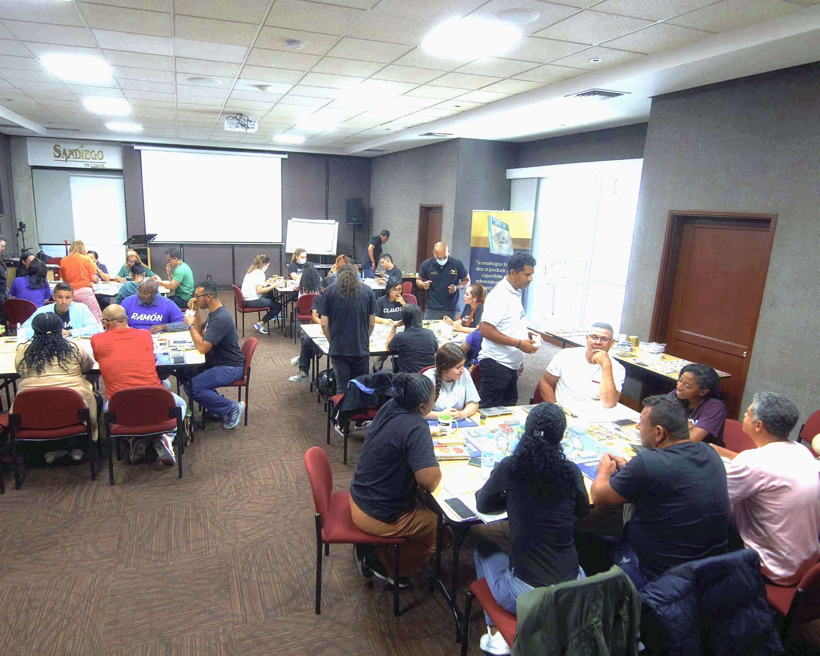Salón lleno durante un entrenamiento: grupos de aprendices juegan el simulador financiero en mesas, acompañados por facilitadores.