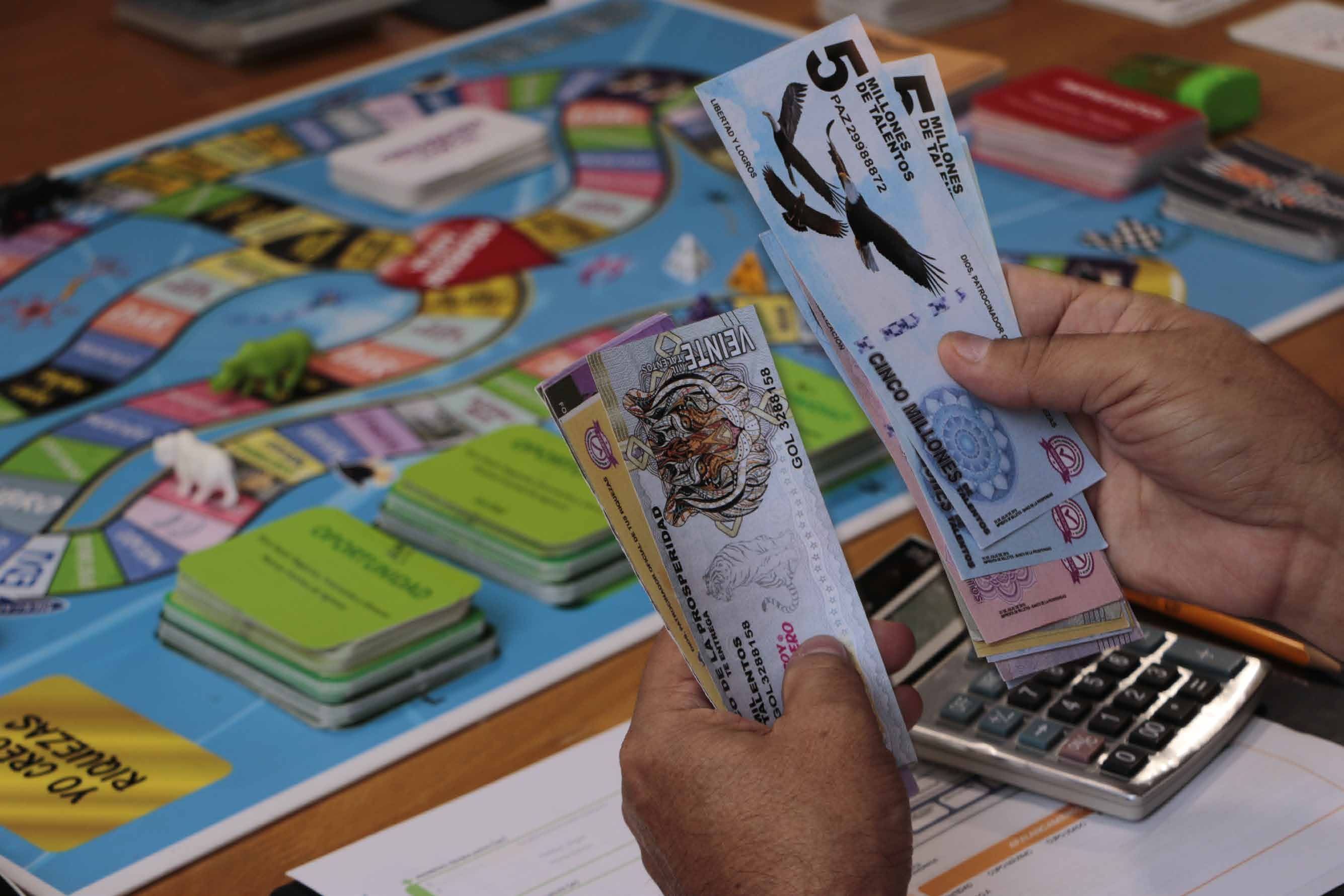 Manos que sostienen billetes de juego del simulador financiero sobre el tablero, junto a una calculadora.